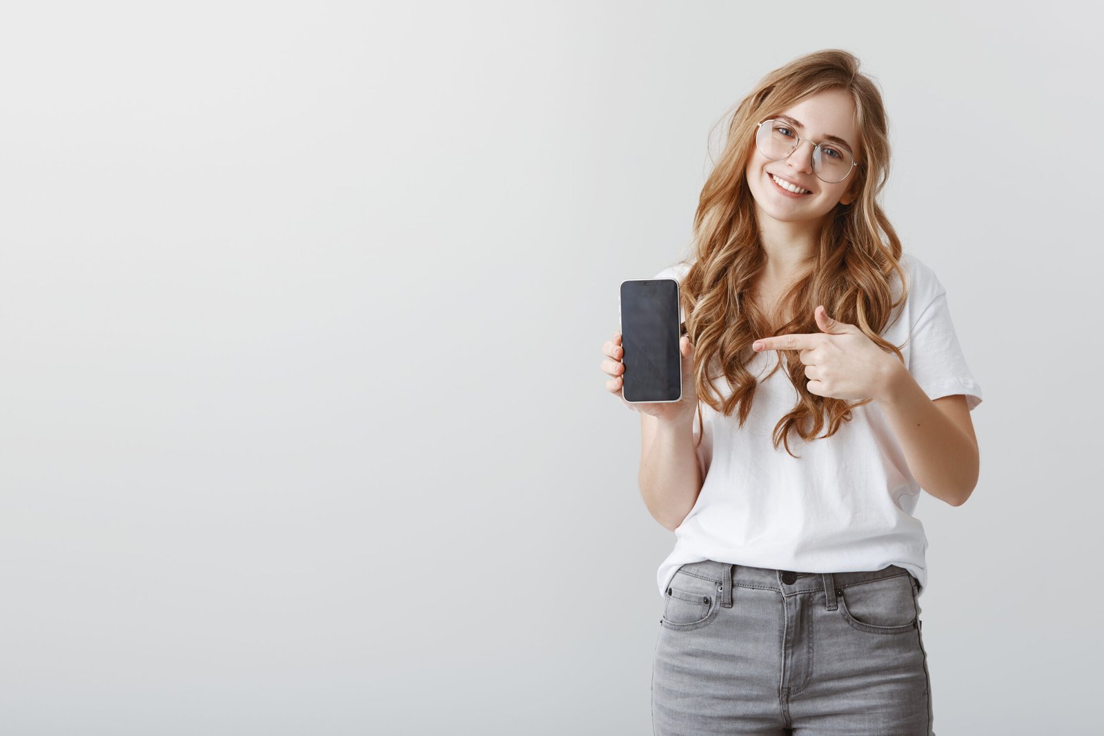 smiling-attractive-blond-girl-glasses-pointing-finger-mobile-phone-screen-showing-application